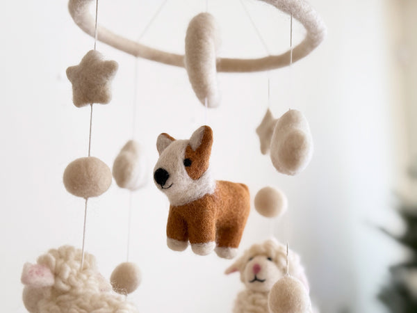 A felt Corgi and Sheep Baby Mobile, suspended from a circular wooden frame, is presented in a close-up shot with a soft, whimsical atmosphere. The mobile features a prominent central brown and white felt corgi, detailed with white markings. Fluffy white wool felt sheep, stars, clouds, and felt balls are visible in the background, all crafted in a neutral palette of soft whites and browns.