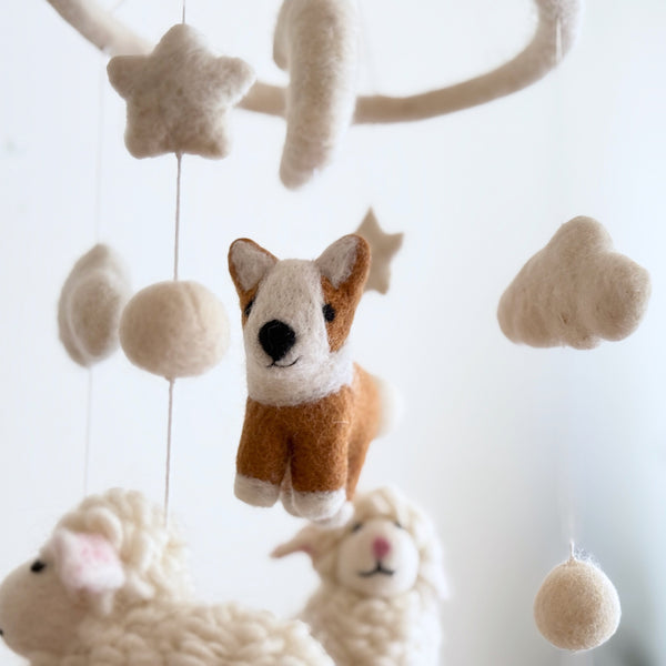 A wool felt Corgi and Sheep Baby Mobile hangs against a bright white background, showcasing a whimsical, neutral-colored nursery decor item. A smiling brown, white, and black wool felt corgi is central, surrounded by fluffy white wool felt sheep, stars, clouds, and felt balls. All elements are suspended from a circular wooden frame, creating a cozy farmhouse aesthetic.