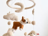 A wool felt Corgi and Sheep Baby Mobile is displayed against a plain white background, creating a soft, calming atmosphere. It features needle-felted figures of a light brown and white corgi, cream-colored sheep with pink inner ears, and matching cream clouds, stars, and felt balls. These charming figures are suspended from a circular wooden frame, crafted from wool felt. The handmade mobile has a whimsical and cozy, rustic aesthetic suitable for a nursery.