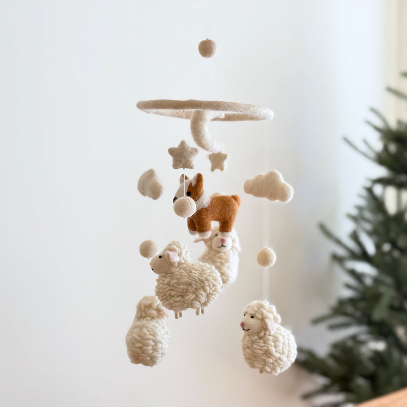 A charming corgi and sheep wool felt baby mobile hangs against a soft white wall, with a green pine tree partially visible on the right, creating a cozy nursery scene. The mobile features needle-felted figures suspended from a circular wooden frame, topped by a beige felt ball. A tan-and-white corgi is encircled by beige stars and clouds, while three fluffy wool felt sheep and more beige felt balls adorn the bottom.