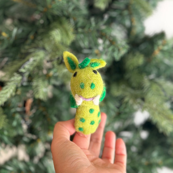 A hand holds a small, green wool-felt dragon finger puppet with a friendly expression and big smile. The dragon has an elongated body with scales, two wings on its back, and whimsical design. It's perfect for storytelling or as a holiday gift for kids.