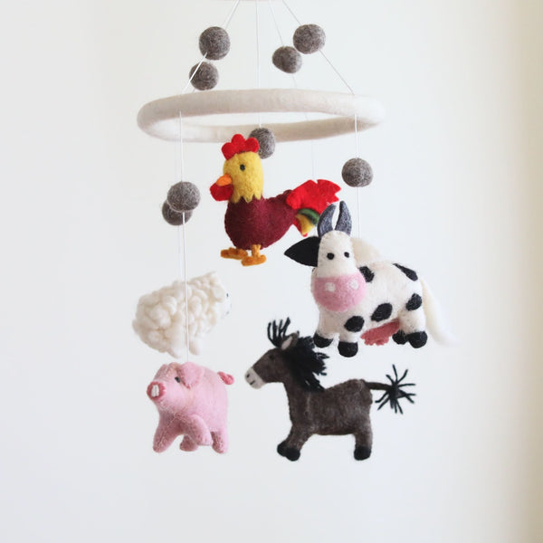 A felt baby mobile, featuring farm animals like a rooster, milk cow, pig, sheep, and donkey, crafted from wool felt with attention to detail, hangs from a white ceiling in a nursery setting.