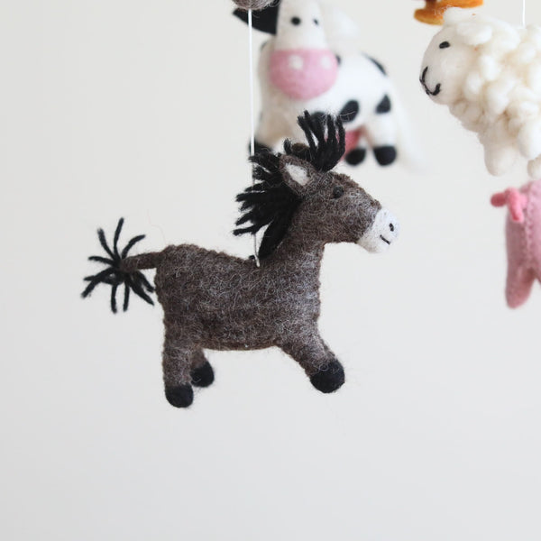 A wool felt baby mobile, 'Farmland Animal Baby Mobile', hangs from a string against a white background. The mobile features various animal figures, including a rooster, milk cow, pig, sheep, and donkey, all made of soft wool felt. Each figure is depicted in a cartoonish style with simple shapes and bright colors, arranged in a circular pattern for balance and harmony.