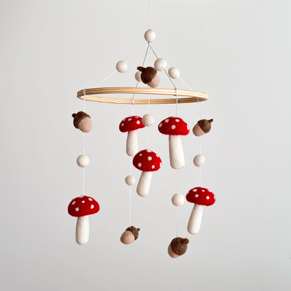 A charming wool felt baby mobile, suspended from a wooden hoop, features handcrafted felt mushrooms and acorns in warm, earthy tones. The natural wool felt decorations add a touch of nature-inspired magic to any room.