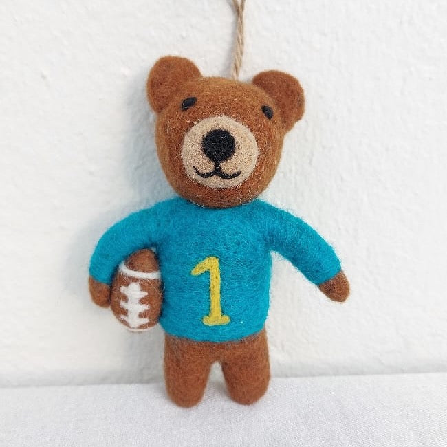 A handcrafted wool felt bear ornament of Joe Bruin, the UCLA football mascot, rests against a clean, bright backdrop. This cheerful, light brown bear ornament wears a blue athletic jersey with a yellow '1' and holds a brown football with white stitching. It features a black nose, eyes, and small, rounded ears. A jute string is attached for hanging this approximately 4.5-inch tall handcrafted felt ornament.