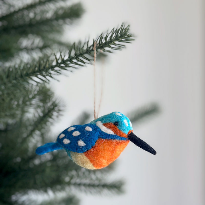 A vibrant, handcrafted needle-felted wool kingfisher ornament hangs from a green Christmas tree branch, evoking festive charm. The colorful felt bird ornament features striking blue and orange with a black beak and small white spots near the tail. A jute string allows it to dangle delicately, perfect for Christmas tree decor.