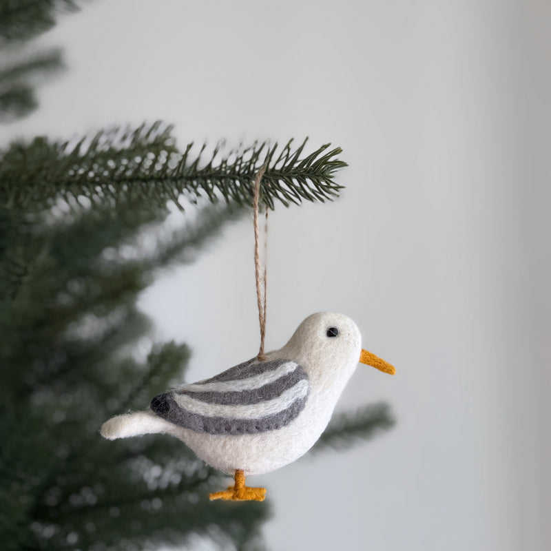A handmade wool felt seagull ornament from the Felt Bird Ornaments set dangles gracefully from an evergreen Christmas tree branch against an off-white background. This distinctive seagull features a predominantly white body with gray striped wings, a small black eye, and bright orange beak and feet. This charming Christmas tree ornament, crafted from wool felt, adds a playful seasonal touch to holiday decor.