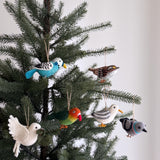 Six handmade wool felt bird ornaments, a blue budgerigar, white throat sparrow, green parrot, grey seagull, white dove, and grey pigeon, are suspended from a festive Christmas tree. Each handcrafted felt ornament features quirky details and wire legs, with the white dove holding a small green leaf. This set adds a playful, whimsical touch against a soft white backdrop.