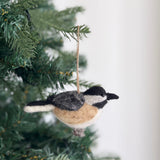 A needle felted chickadee ornament hangs from a green Christmas tree. This handcrafted wool felt bird ornament features a black head, a grey and white patterned back, and a tan chest. A jute string loop is attached for easy hanging, adding a whimsical touch to the festive decor.