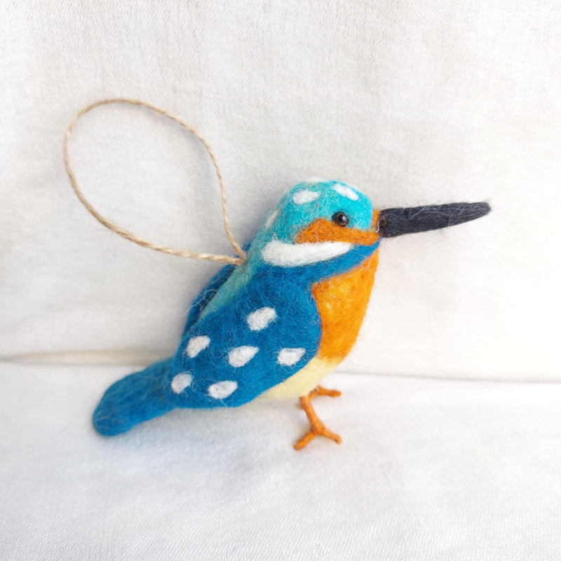 A single felt kingfisher ornament from a set of six felt bird ornaments is displayed against a clean white backdrop. The vibrant kingfisher features a blue head dotted with white, an orange chest, and complementary white and blue wings and tail. Its lifelike appearance is enhanced by a long black beak and delicate orange wire legs. A jute string transforms this unique Christmas ornament into a delightful hanging decoration.