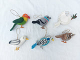 Six wool felt bird ornaments are displayed against a bright white fabric backdrop. The charming set includes a vibrant green parrot, a gray pigeon, a white dove carrying a twig, a white and gray seagull, a light blue budgerigar, and a brown and white sparrow. Each handcrafted felt ornament features a jute string for easy hanging.