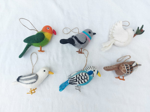 Six wool felt bird ornaments are displayed against a bright white fabric backdrop. The charming set includes a vibrant green parrot, a gray pigeon, a white dove carrying a twig, a white and gray seagull, a light blue budgerigar, and a brown and white sparrow. Each handcrafted felt ornament features a jute string for easy hanging.
