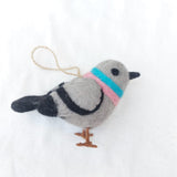 A charming wool felt pigeon ornament is prominently displayed against a bright white background. This handmade Christmas tree ornament features a soft gray body with black tail markings and black wire legs. A blue and pink band encircles its neck, while black eyes and beak give it an endearing expression. A jute string is attached, ready for hanging.