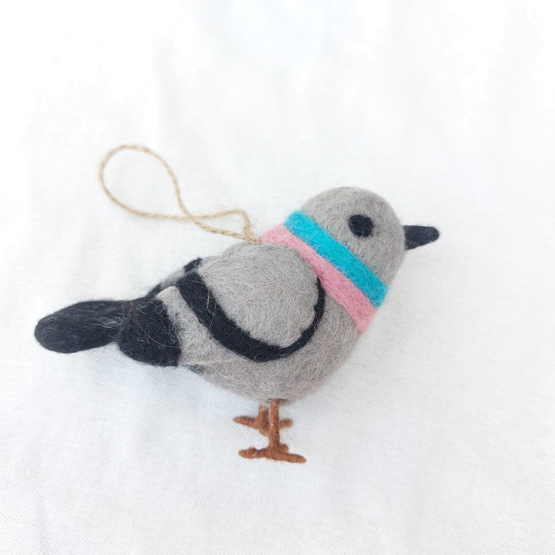A charming wool felt pigeon ornament is prominently displayed against a bright white background. This handmade Christmas tree ornament features a soft gray body with black tail markings and black wire legs. A blue and pink band encircles its neck, while black eyes and beak give it an endearing expression. A jute string is attached, ready for hanging.