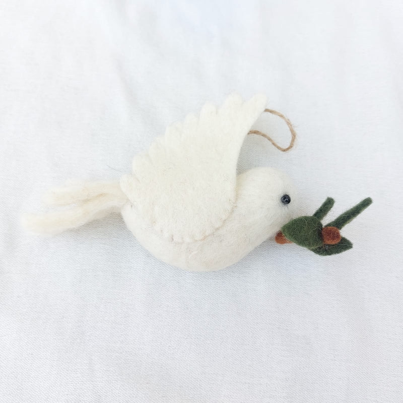 A handcrafted wool felt dove ornament, part of a set of felt bird ornaments, is displayed on a textured white surface. This creamy white dove features a small black eye and holds a green branch with brown berries in its beak, exuding a soft, calming charm. A jute string is attached for hanging.