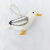 A wool felt seagull ornament, one of the handmade felt bird ornaments, is displayed against a plain white background. This charming seagull has a white body, a yellow beak and legs, and gray and white striped wings with black tips. A brown jute string loop is attached at the top, ready for hanging on a Christmas tree.