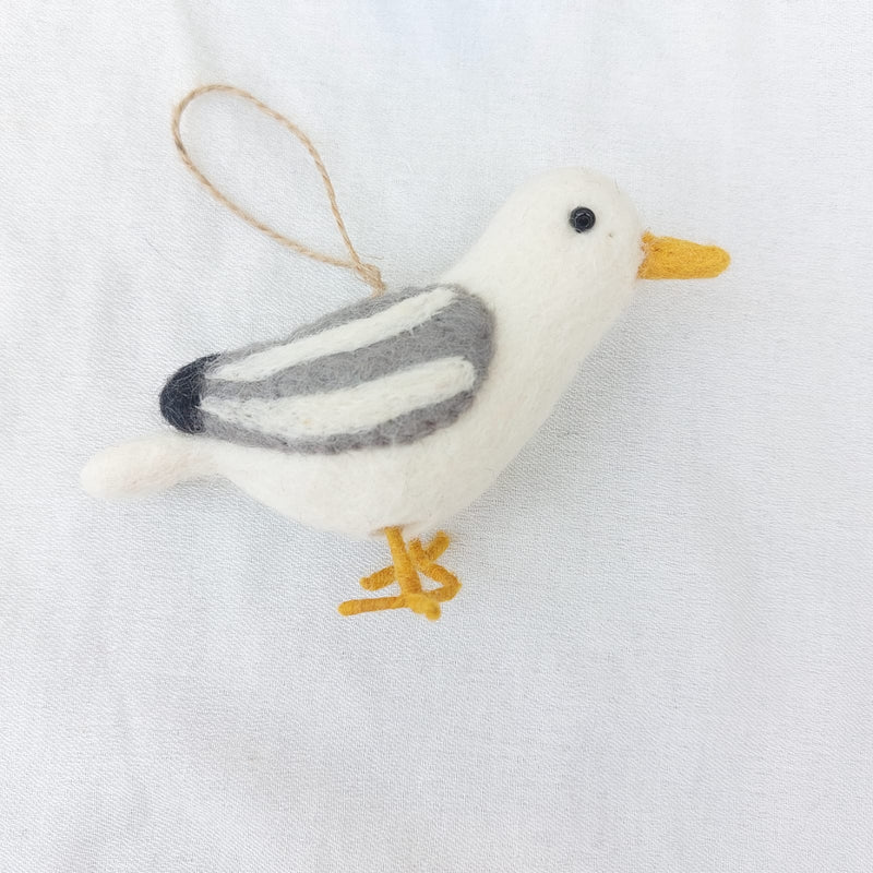 A wool felt seagull ornament, one of the handmade felt bird ornaments, is displayed against a plain white background. This charming seagull has a white body, a yellow beak and legs, and gray and white striped wings with black tips. A brown jute string loop is attached at the top, ready for hanging on a Christmas tree.