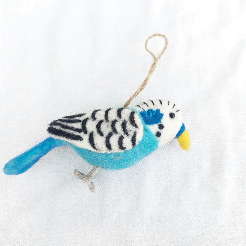 A cheerful 100% wool felt blue budgerigar ornament is displayed against a bright white textured background. This charming felt bird features a dual-tone blue body, a white head with black curved markings, a yellow beak, and small black eyes. It includes light gray wire legs and a jute string for easy hanging.