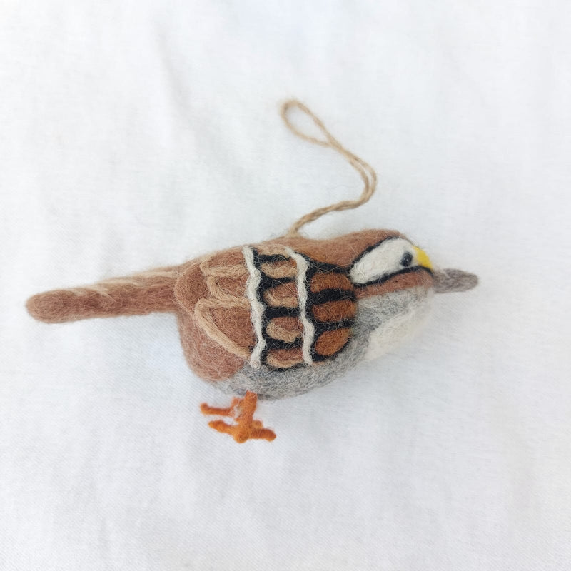 One wool felt white throat sparrow ornament is presented against a clean white background, showcasing its handmade charm. This felt sparrow ornament features natural browns, white, black, and gray plumage, with delicate wire legs and a jute string for hanging. Its soft, slightly fuzzy texture highlights its unique appeal.