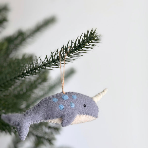 A Felt Blue Narwhal Ornament hangs on a Christmas tree branch, showcased against a plain white backdrop. This handcrafted felt narwhal ornament features a soft gray wool felt body adorned with playful blue spots and a prominent stitched ivory tusk, complete with a jute string for hanging.