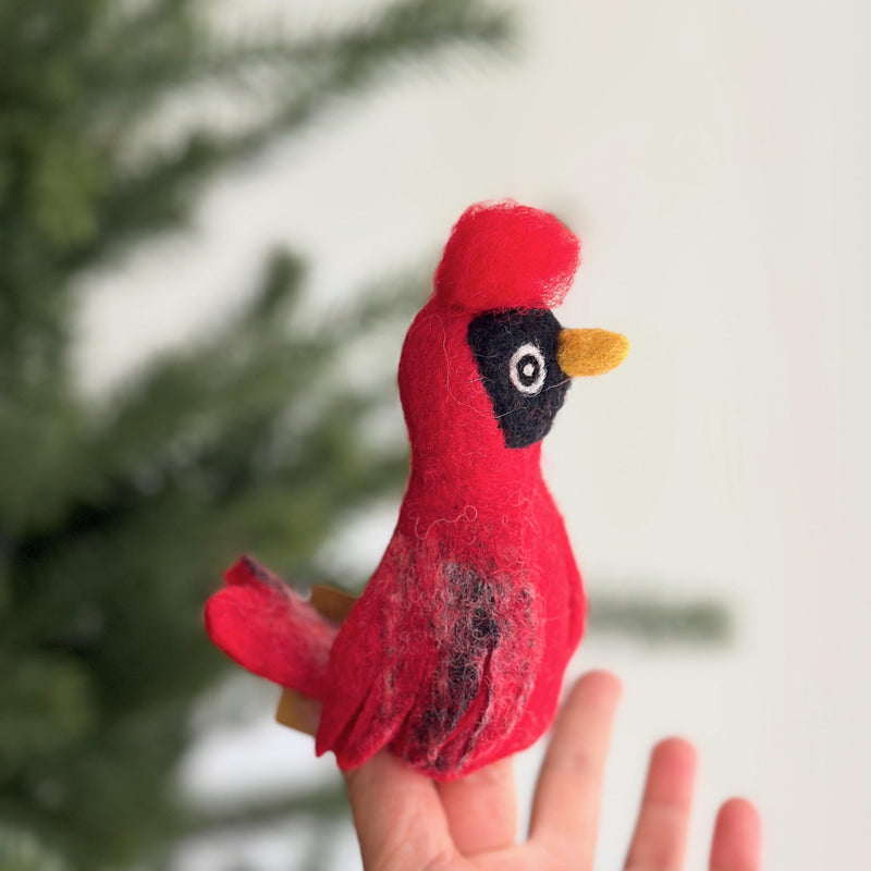 A needle-felted blue jay finger puppet is held gently in a female hand, set against a blurred background of a Christmas pine tree and a white wall. This handcrafted blue jay puppet showcases striking blue felt on its head, wings, body, and tail, accented with a distinctive crest and textural variations suggesting feathers. Its detailed craftsmanship supports fair employment for Nepali artisans.