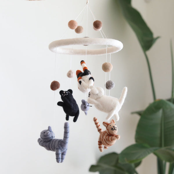 A felt cat baby mobile hangs from a ceiling, featuring five adorable kittens in various colors and patterns. The background is minimalistic with a white wall and a plant on the left side. The mobile showcases intricate hand-stitched details, with each kitten uniquely colored: white, black, gray, tabby, orange tabby, and calico.