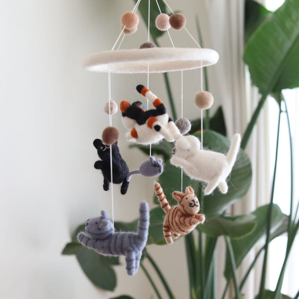 A Felt Cat Baby Mobile hangs from a white string against a white wall, featuring five colorful kittens: white, black, gray, tabby, orange tabby, and calico. Each kitten has a unique facial expression and pose, giving the impression of playful movement.