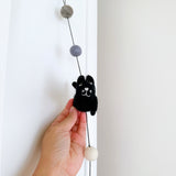 A charming felt cat garland is gently held against a stark white door and wall. The prominent black wool felt cat, with its three-dimensional design and embroidered smiling face with whiskers, is featured in the center. It connects to a string of light gray, dark gray, and white felt balls, with a copper bell at the bottom.