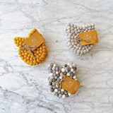 Three sets of handcrafted wool felt cat coasters are displayed on a marbled surface. Each coaster is approximately 4 inches in diameter and 0.5 inches thick, made from tightly arranged felt balls with subtle cat ear details. The sets come in mustard yellow, mixed gray, and solid light gray. Each set includes four coasters, bundled with jute twine and a branded tag that highlights their artisanal charm.