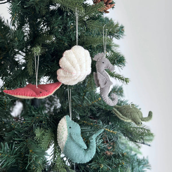 A set of five wool felt sea animal Christmas ornaments hangs festively on a green Christmas tree. This handmade collection includes a red starfish, a white shell with small white dots, a gray seahorse, a green sea turtle, and a teal whale with a white underbelly. Each handcrafted felt ornament has an endearing texture and is suspended by a delicate white cotton string.