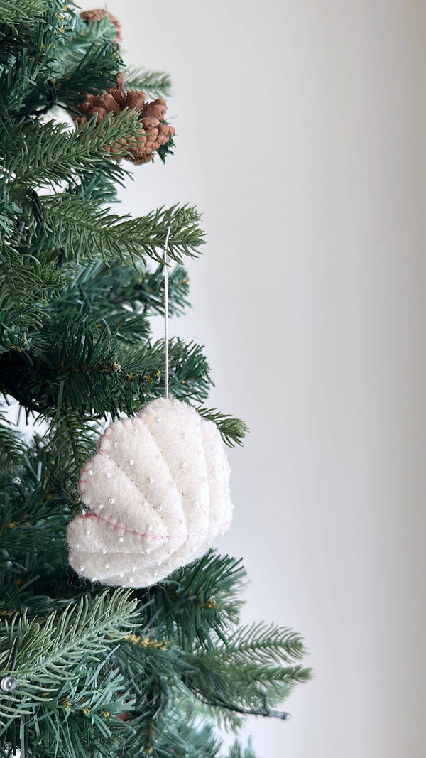 A white wool felt seashell ornament, part of a sea animal felt ornament set, is gracefully suspended from a green Christmas tree branch. This handcrafted felt ornament features a textured surface embellished with tiny white beads. It hangs amidst green pine needles and brown pinecones, evoking a simple, warm, and inviting holiday decor.
