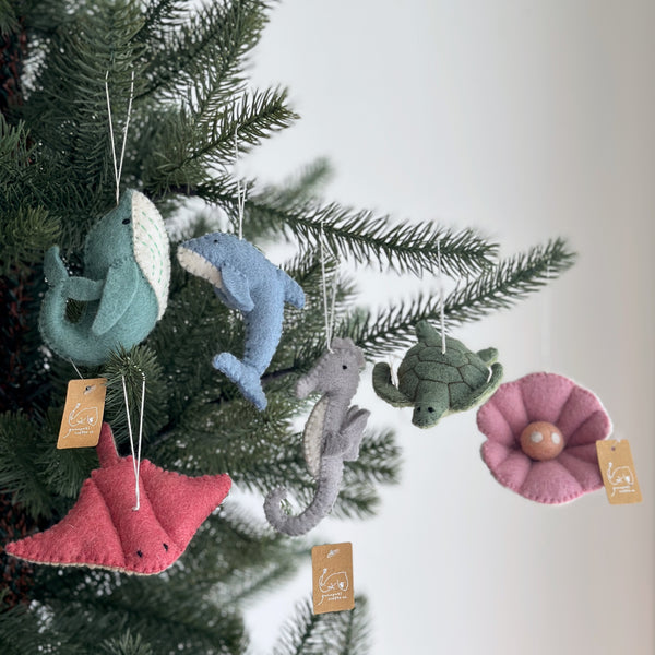 A set of six unique felt ocean creature Christmas ornaments hangs from a festive Christmas tree against a soft, neutral backdrop. The handmade wool felt ornaments include a light blue shark, a light blue streamlined dolphin, a gray textured seahorse, a dark green patterned turtle, a red flat stingray, and a pink pearl. White cotton strings secure each ornament, which also features a small cardboard tag.