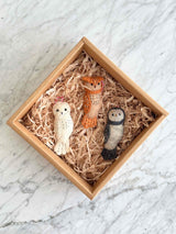 A set of three hand-stitched felt owl ornaments are nestled in a square cardboard box filled with shredded light-brown paper, resting on a bright, subtly marbled surface. The set includes a white, an orange, and a gray owl, each featuring round black eyes, stitched details, and a red and white cotton string for hanging.