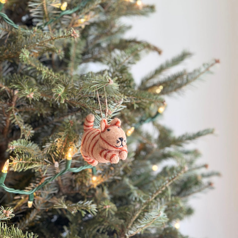 A handcrafted felt cat Christmas ornament, part of a set of four, hangs gracefully on a branch of a festive, lit Christmas tree. The light brown felt cat features darker brown stripes, rounded ears, a short tail, and a stitched black nose and mouth, ready for display.