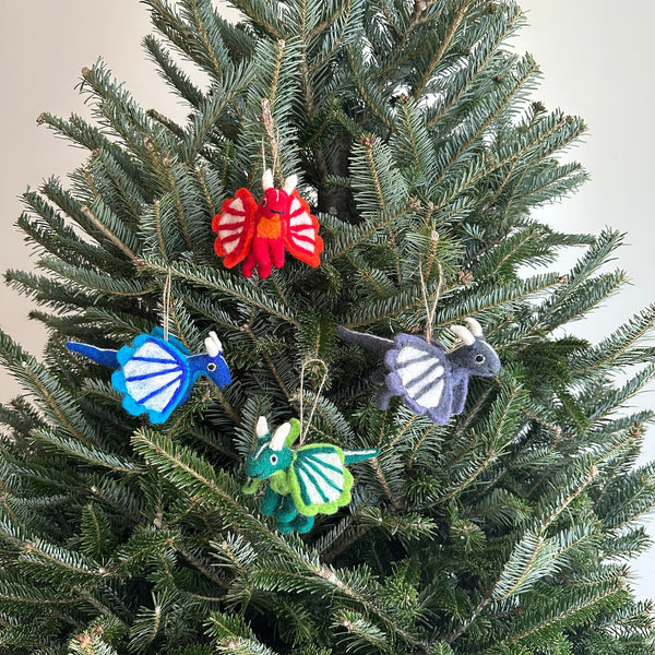 A set of four vibrant felt dragon Christmas ornaments hangs from the branches of a lush Christmas tree. Each handcrafted felt dragon ornament, suspended by a jute string, comes in red, blue, green, or grey. The red and grey dragons feature white wing accents, the blue a light blue, and the green a light green, all with tails and horns.