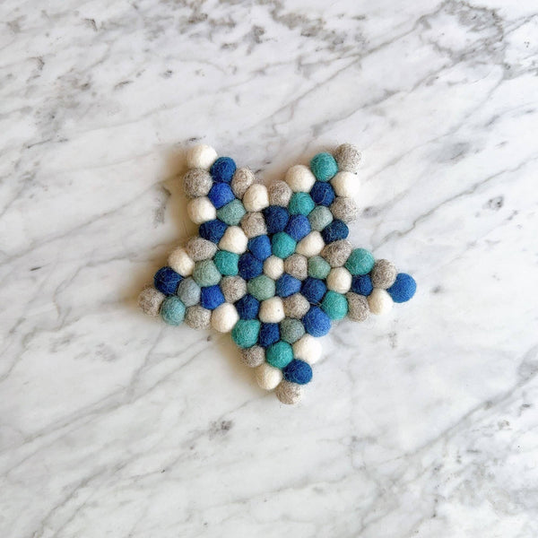 A blue star felt coaster is displayed on a marble surface. This handcrafted coaster is made from individual hand-stitched felt balls in shades of white, gray, and blue, offering a charming and unique design suitable for home decor.