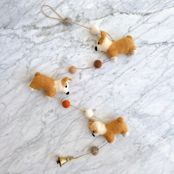 The Felt Corgi Garland is displayed diagonally on a white marble surface. It features three light brown felt corgis with white markings on their faces and paws. Small, round felt balls in white, brown, and orange are placed between each corgi, with a twine loop at one end.
