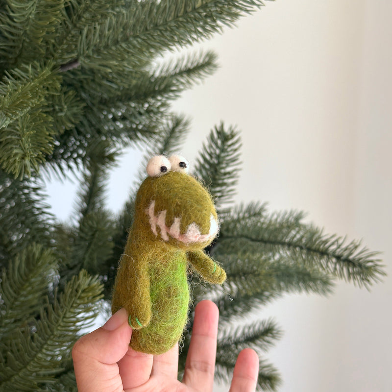 A hand holds a small, green felt gator finger puppet, positioned in front of a Christmas tree. The overall mood is playful and whimsical, evoking holiday cheer and childhood imagination.