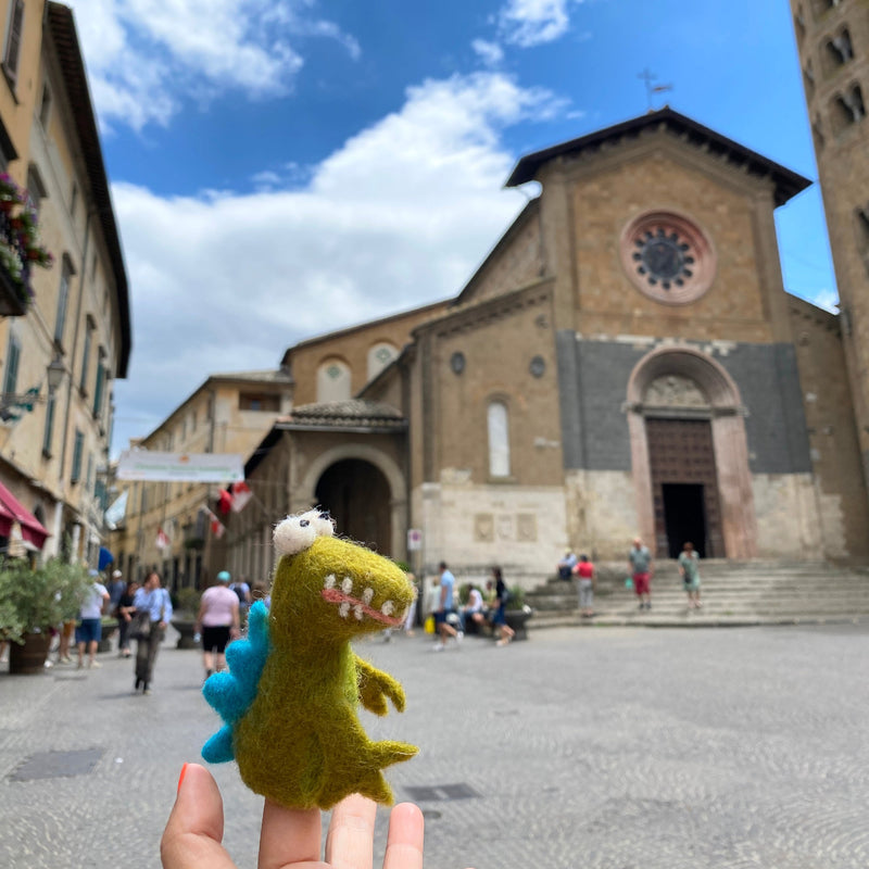 A hand holds a small, green felt gator finger puppet in front of a charming church with a clock tower on a cobblestone street. The background features people walking on the sidewalk, creating a playful and whimsical atmosphere. This adorable wool felt finger puppet is designed to bring stories to life during imaginative playtime.