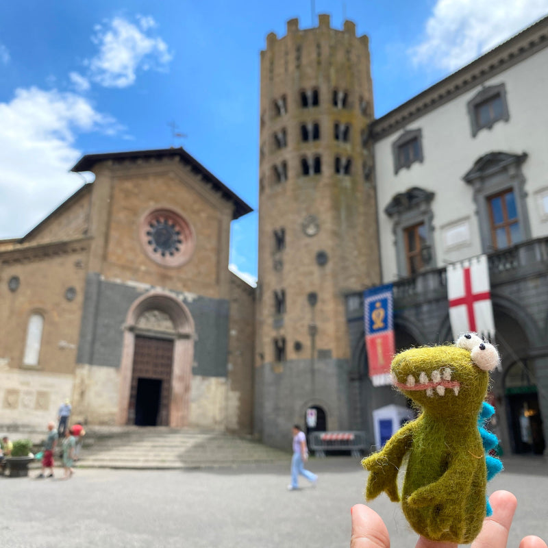 A hand holds a small, green felt alligator finger puppet in front of a stone church with a tall tower. The puppet has a wide smile and is positioned on a table or surface, drawing attention due to its vibrant color and playful design. The church provides a contrasting backdrop to the whimsical nature of the puppet. This felt alligator finger puppet features a cute and whimsical gator design, crafted from high-quality wool felt with keen attention to detail.