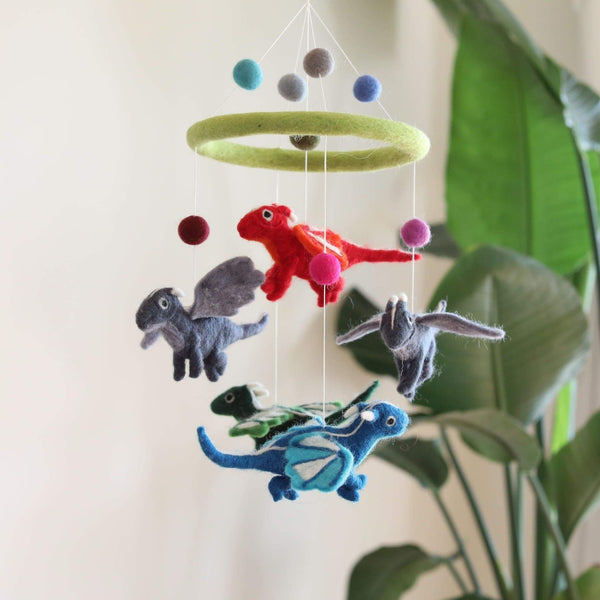 A baby mobile hangs from the ceiling, featuring four felt dragons in various colors and patterns against a white wall. The mobile's central frame features a ring of small spheres, partially obscured by a plant in the foreground.