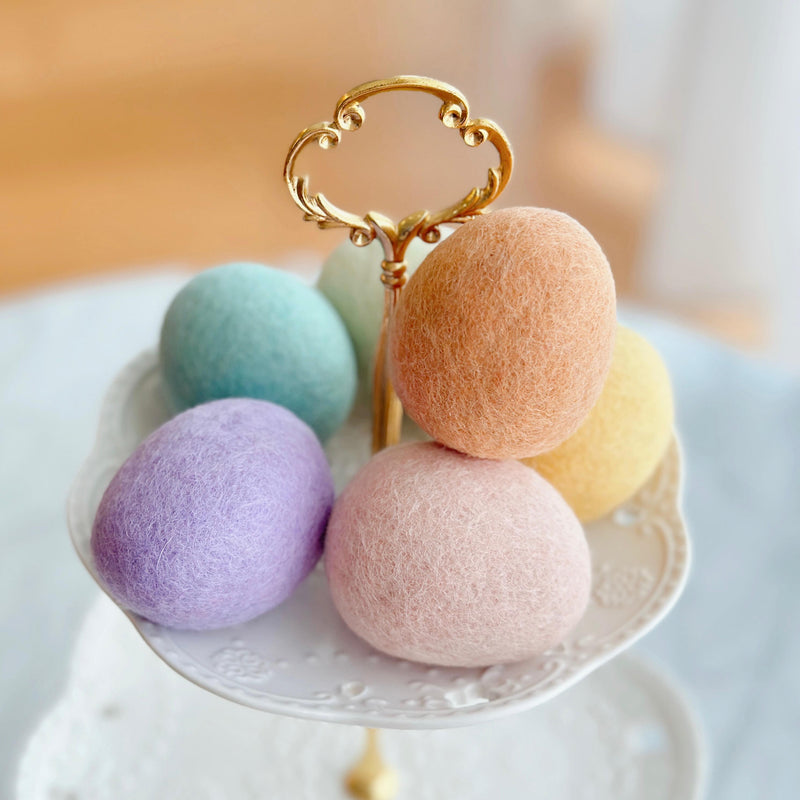 Six life-sized wool felt Easter eggs are elegantly displayed on the top tier of a white, two-tiered decorative stand with a gold handle. The pastel felt Easter eggs include shades of light blue, peach, yellow, pink, green, and lavender. They are densely arranged, filling the stand's top tier, which features a delicate, floral-patterned rim, evoking a dreamy springtime charm.