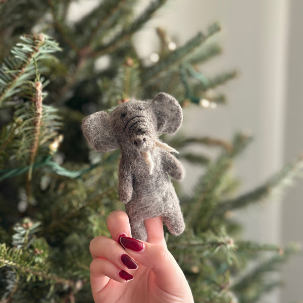 A charming wool felt elephant finger puppet, crafted by hand using 100% wool, rests on a festive Christmas tree background. The intricate textures of the puppet mimic an elephant's skin, with a wire attached to its nose for added realism. This handmade felted finger puppet features a unique design and is made from needle-felted wool felt.