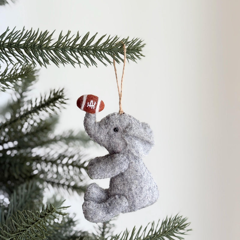 A gray wool felt elephant Christmas ornament, playful and handcrafted, hangs from a festive Christmas tree branch. The charming elephant is seated, with front legs slightly raised, and its trunk holds a brown football with white stripes. A small black eye and a jute string for hanging complete this whimsical, unique Christmas ornament.