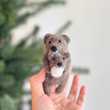 A handmade American river otter felt finger puppet is gently held by a female hand against a soft, diffused background with a hint of green foliage. This charming felt finger puppet features a mother otter cradling her baby, crafted from textured brown felt. The baby otter has a white belly, and both otters have tiny black eyes and noses, creating a sweet, endearing appearance.