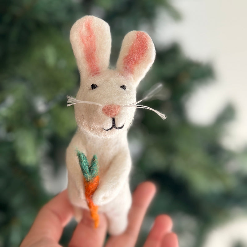 A hand holds a small wool felt white rabbit finger puppet against a blurred indoor setting with Christmas decorations. The bunny has long ears, a white face with black eyes, and is holding a carrot in its paws. This handmade felt finger puppet features a cute and cuddly design, ideal for imaginative play or storytelling.