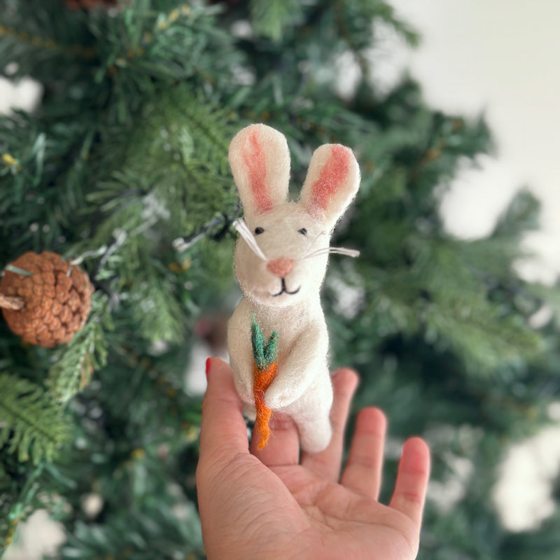 A hand holds a small wool felt white rabbit finger puppet with white inner ears, long whiskers, and holding a carrot. The background features a Christmas tree with pine cones and decorations.