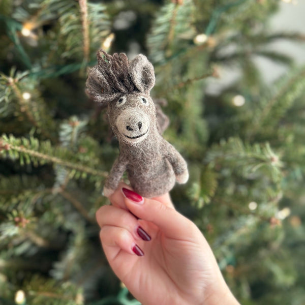 A handmade wool felt donkey finger puppet, resting on a person's hand against the backdrop of a festive Christmas tree. The gray donkey features a smiling face and stands upright with its legs spread apart. Made from 100% wool felt, this unique finger puppet is perfect for storytelling.