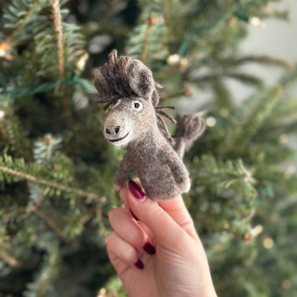 A handmade wool felt donkey finger puppet, resting on a person's hand, is showcased against the festive backdrop of a Christmas tree. The donkey features a friendly expression with a big smile, looking directly at the camera. Made from 100% wool felt material, it showcases visible stitching details on its body and legs.