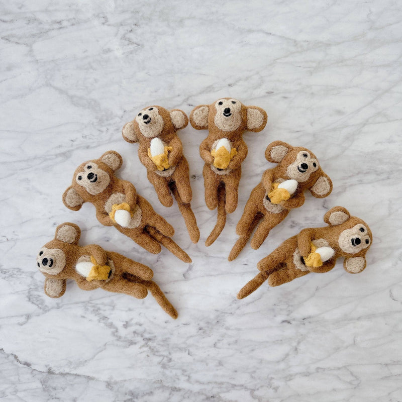 Multiple handcrafted wool felt monkey finger puppets are playfully arranged on a white and gray marble surface. Each warm brown felt monkey finger puppet smiles broadly while holding a small, freshly peeled banana. The visible texture highlights their unique, handmade craftsmanship, perfect for imaginative play.